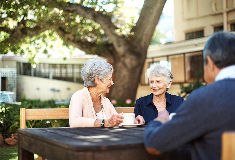 Two residents with coffee are sitting next to each other at a table smiling at each other. Another resident is sitting across from them. 