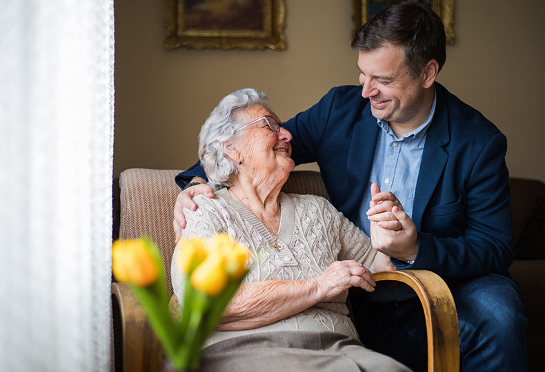A resident is sitting down smiling at a person holding hands. There are yellow tulips in the room.