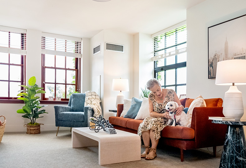 resident with dog in living room with sun shining thru windows.
