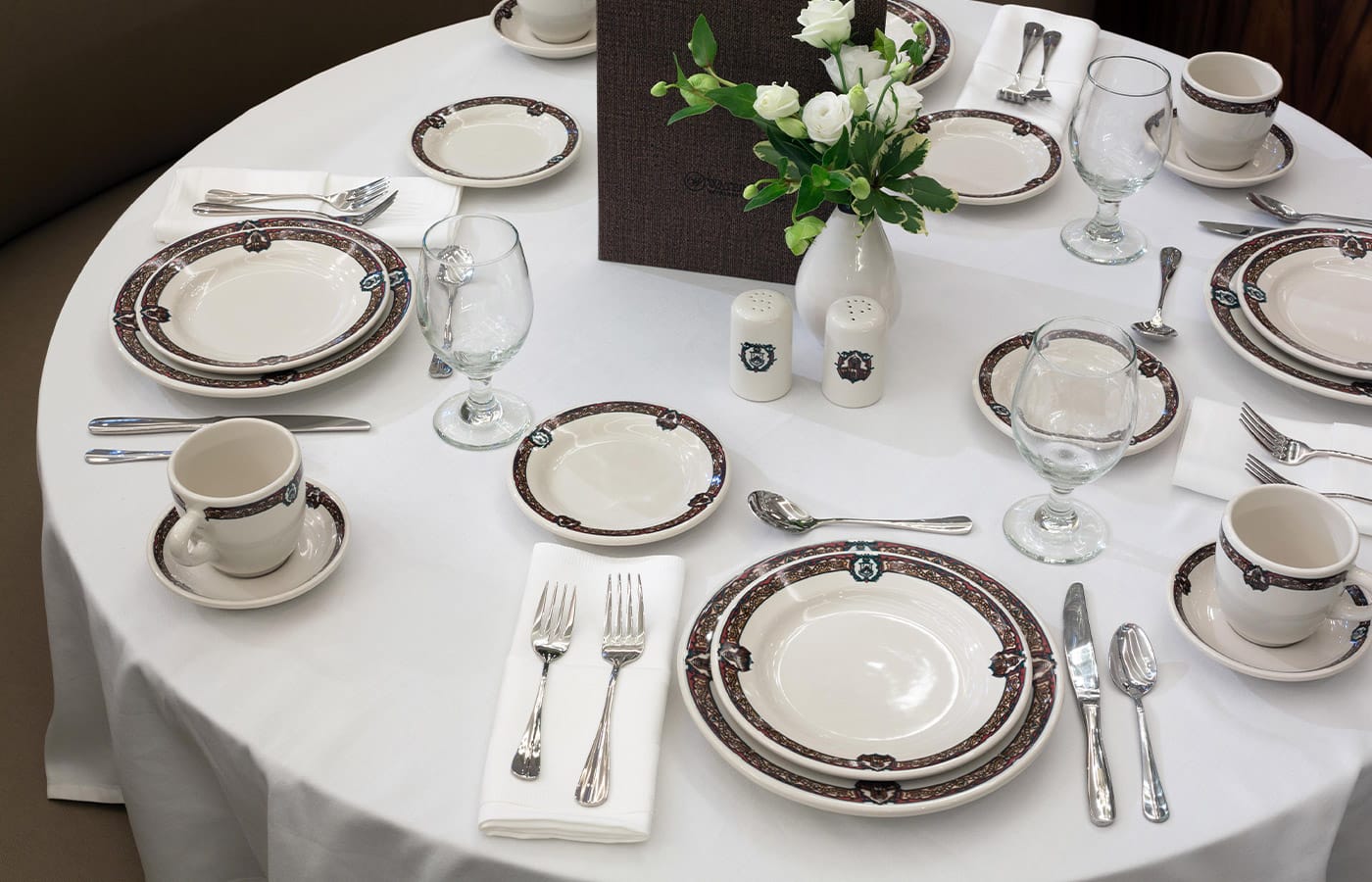 A table set for five with white linens and fine china and accompanying cutlery around a white floral centerpiece.