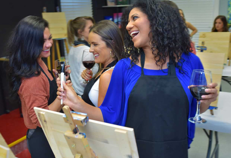 A few people laughing and enjoying wine during a paint class.