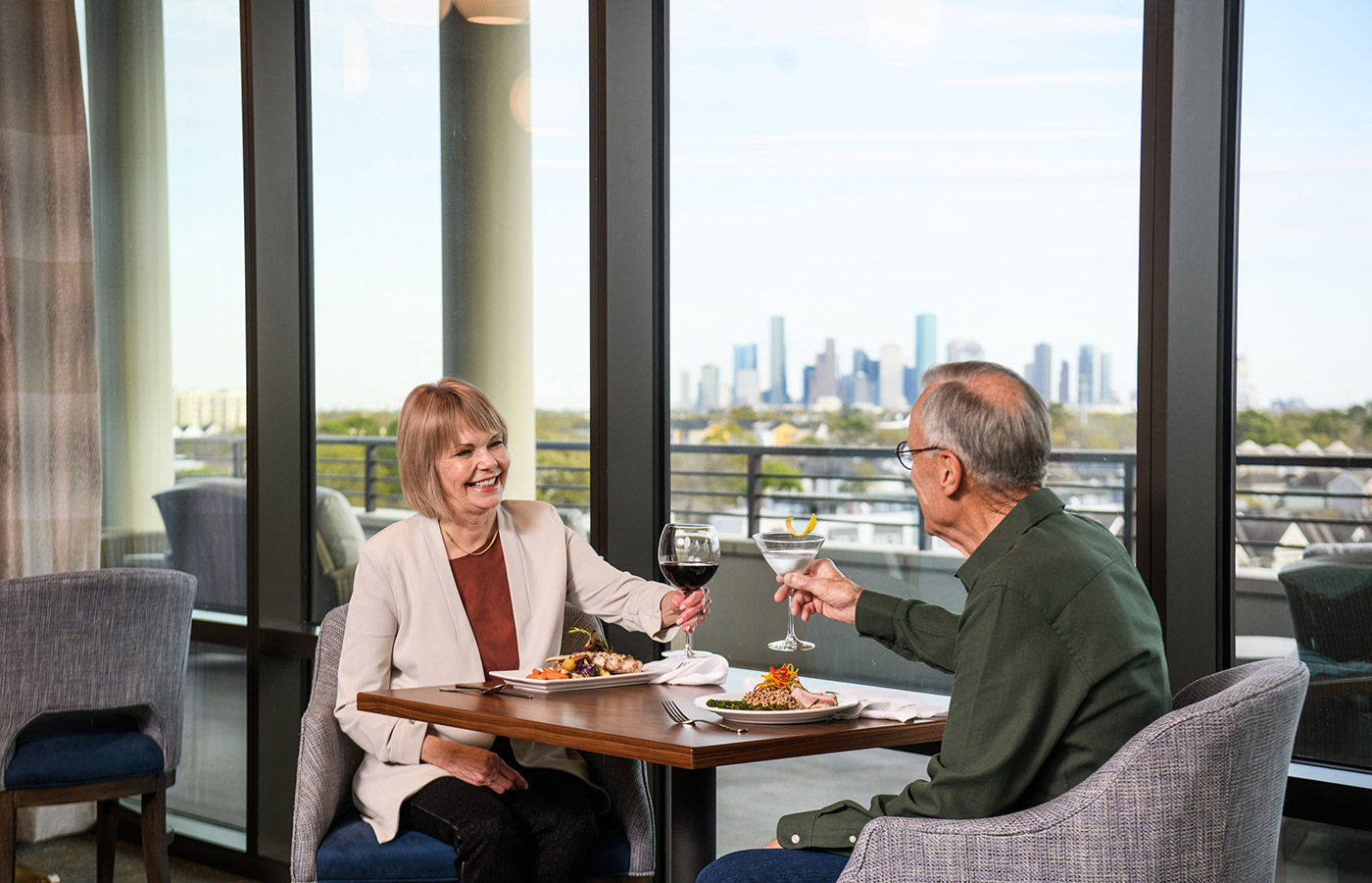 Two residents are sitting by a large window in a dining area enjoying cocktails.