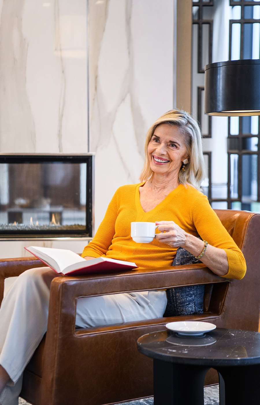 A resident is sitting in front of a fire place and reading a book with a cup of coffee.