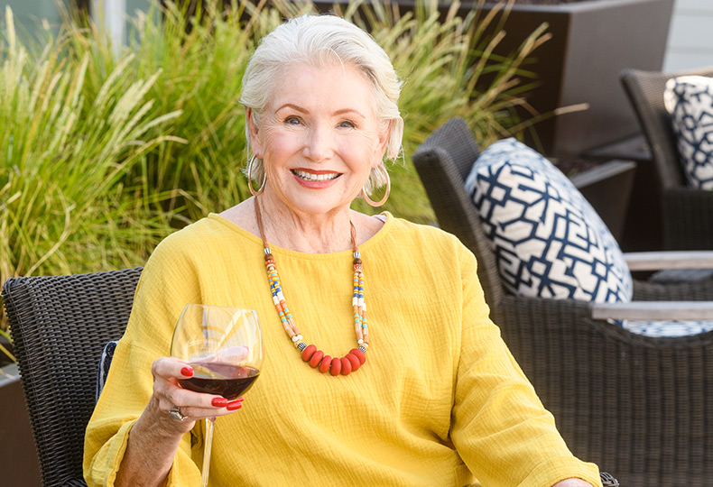 A resident is smiling and sitting outside on the patio area while holding a glass of wine.