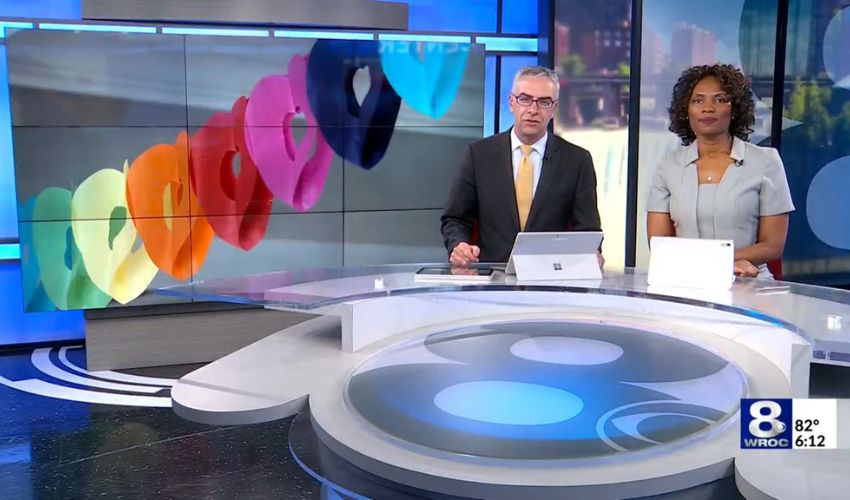 A news crew sitting at a desk with a pride banner behind them.
