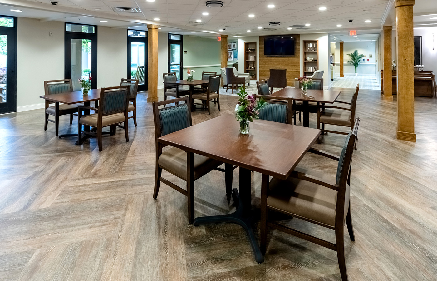 The memory care activity room with tables, chairs and a tv on the wall.