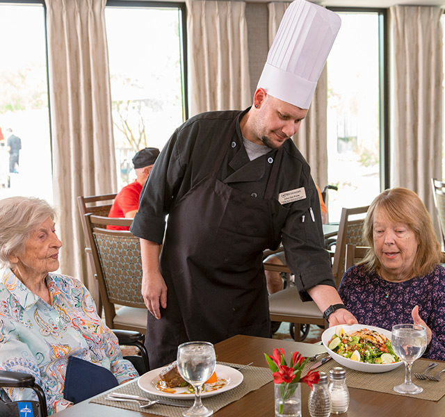 A chef is placing a plate of food in front of a resident in a dining area.