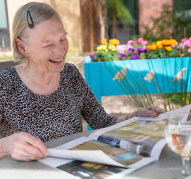 A resident sitting outside at a table smiling and reading a magazine
