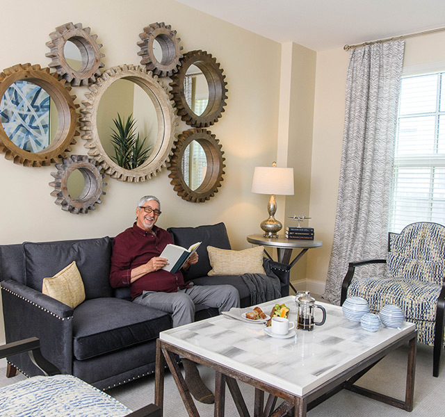 Smiling resident sitting on couch holding a book in large decorated living room.
