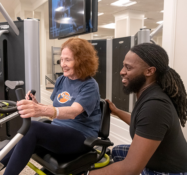 resident on fitness machine in gym with a team member kneeling down and assisting