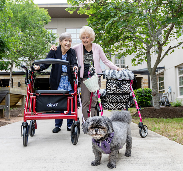 Two residents with walkers walking a dog in the garden.