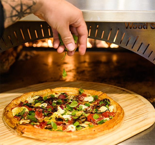 Someone adding toppings to a pizza near a wood fire oven.