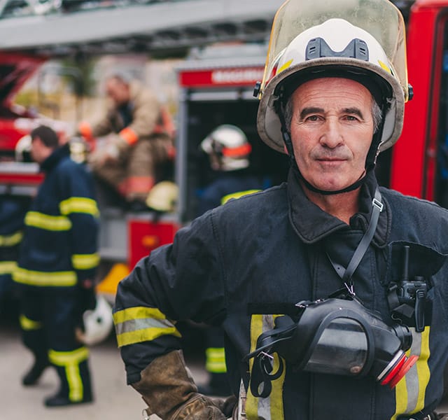 A fire fighter standing in full uniform in front of a fire truck while other crew members stand in a circle talking