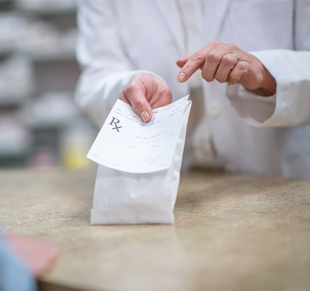 A pharmacist in a white lab coat holds out a processed and packaged prescription.