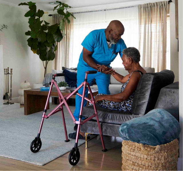 A caregiver gently assists an older adult in a comfortable living room.