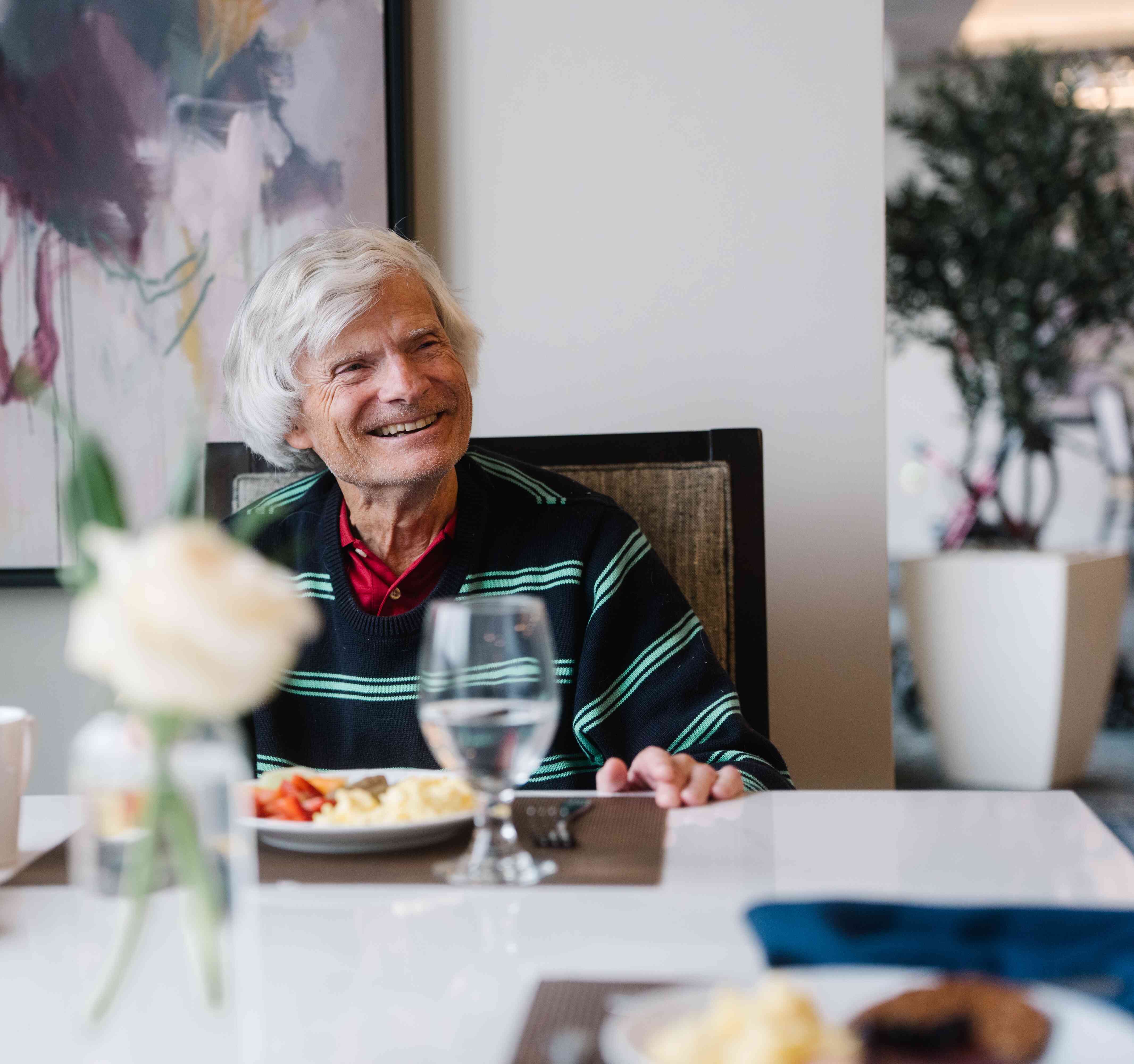 A resident smiles across the dining table while enjoying a meal in the community dining room.