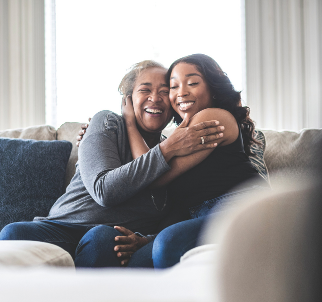 Older woman and younger woman hugging and smiling while sitting on a couch.