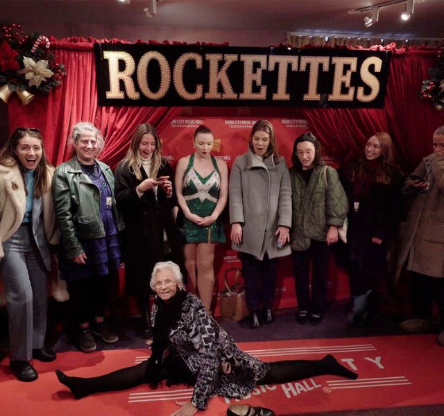 Resident doing the splits in front of the Rockettes step and repeat wall.