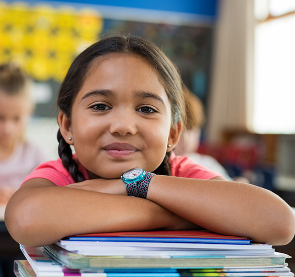 A child is in school resting their arms and head on a pile of books.