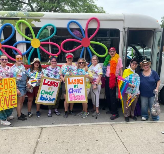 Shuttle with residents from community standing in front of it. Everyone is dressed in bright colors for Pride parade.