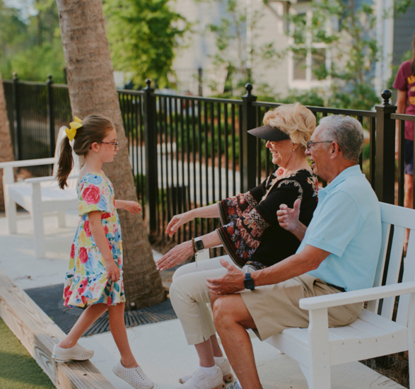 Grandparents sitting on a bench. Grandchild in bright dress talking to them.
