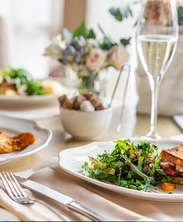 Accordion image Elegant table setting showing three plates of breakfast-type food - an omelet, a salad, and a sandwich. White linens, sparkling water and pink flowers also adorn the table.