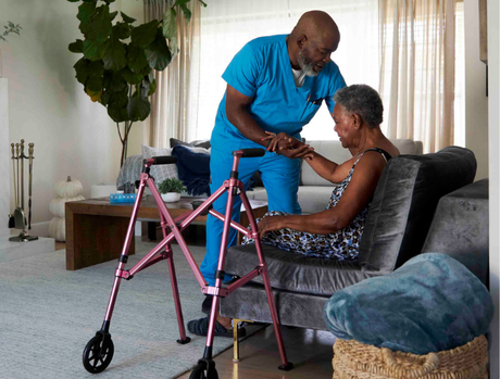 A caregiver gently assists an older adult in a comfortable living room.