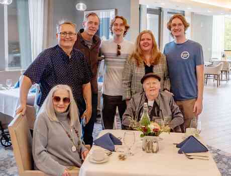 A family gathers around a resident in the dining room, sharing a warm moment across generations in a bright, welcoming setting.