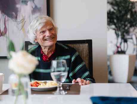 A resident smiles across the dining table while enjoying a meal in the community dining room.