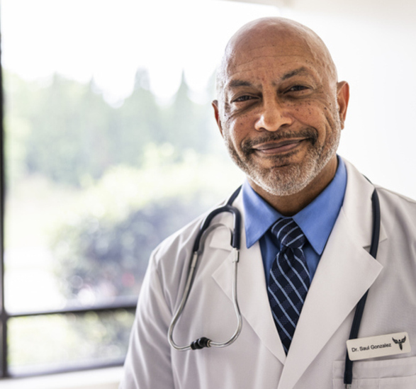 Doctor in white coat standing in hospital lobby smiling at camera.
