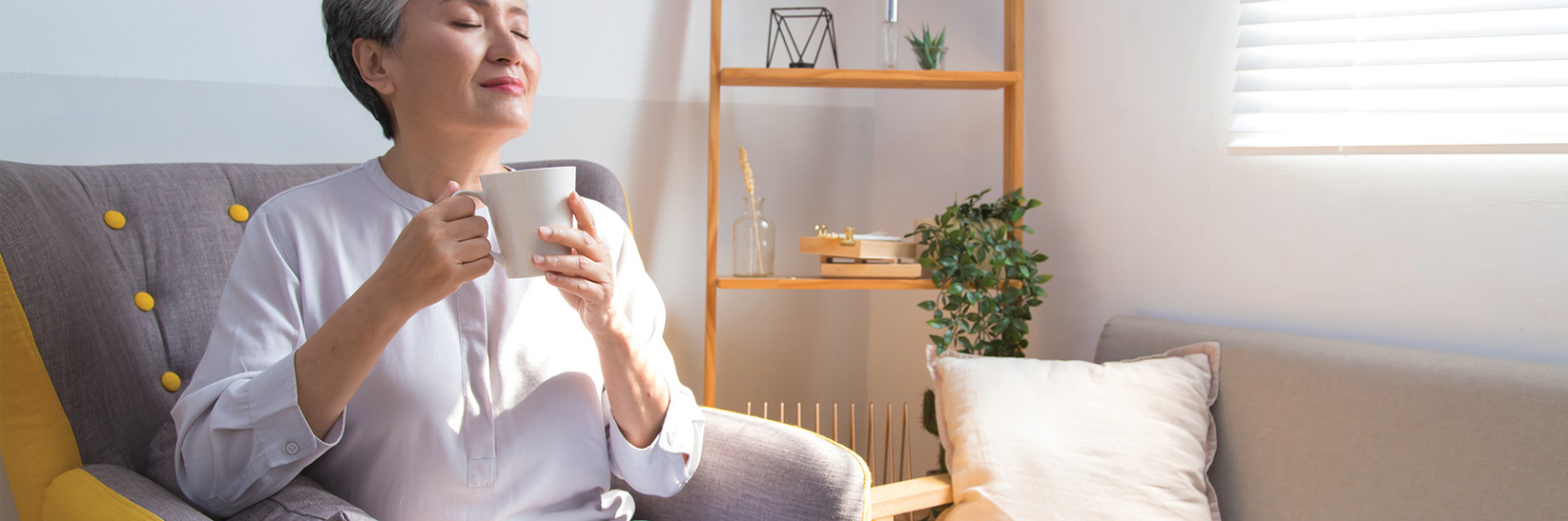 Person with eyes closed, sitting in chair enjoying cup of coffee with sunlight coming in through nearby window. Person with eyes closed, sitting in chair enjoying cup of coffee with sunlight coming in through nearby window.