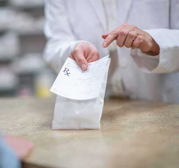 A pharmacist in a white lab coat holds out a processed and packaged prescription.