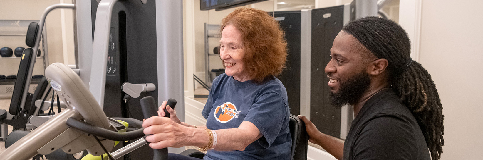 A caregiver is assisting a resident in a fitness center. A caregiver is assisting a resident in a fitness center.