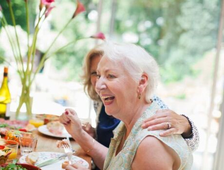 Two people are smiling while eating a meal. One has their arm around the other.