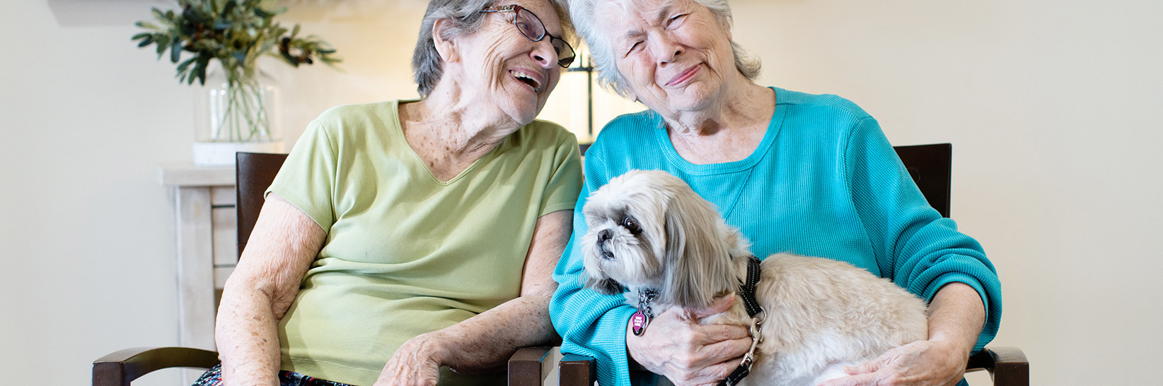 Two residents sitting down holding a dog. Two residents sitting down holding a dog.