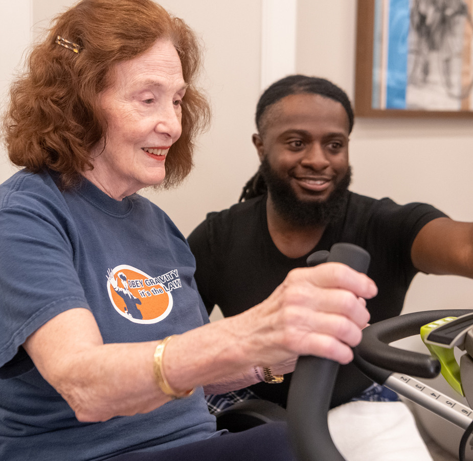 A resident is on a fitness machine with assistance from a caregiver.