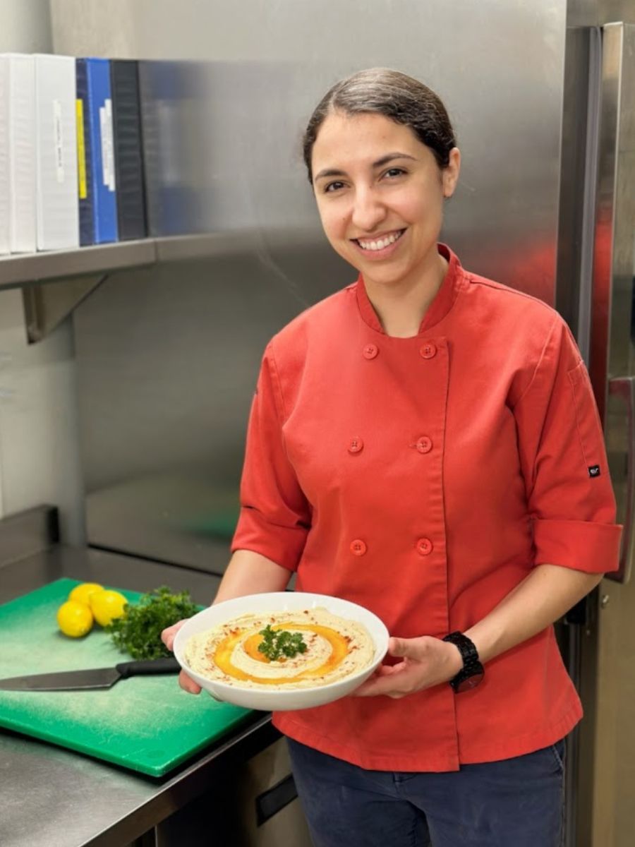 Salwa, a chef, holding a dish of hummus in a kitchen.