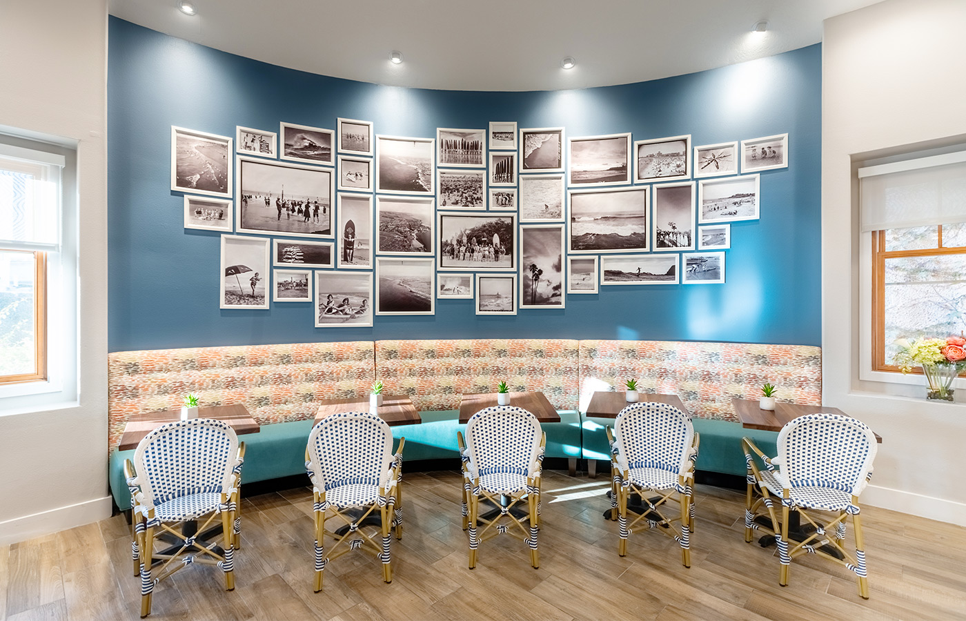 wall in cafe that has multiple black and white beach photos and tables alongside a large booth with woven chairs on the other side