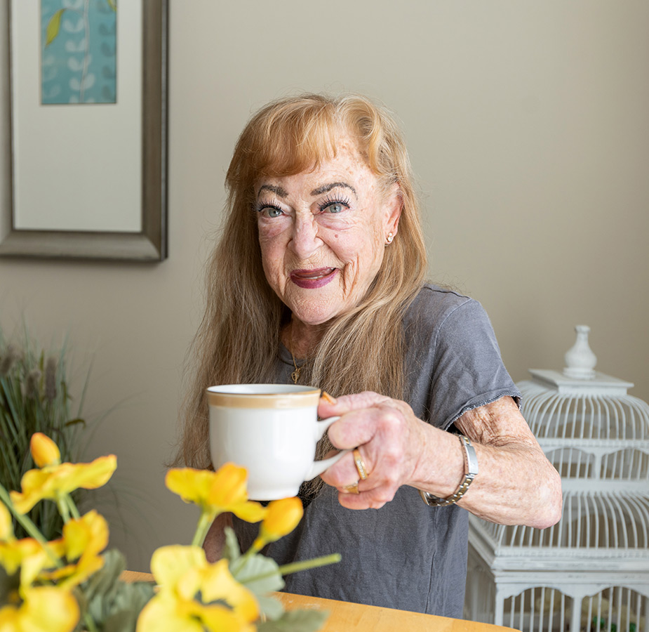 A resident lifting her cup of coffee while smiling into the camera.