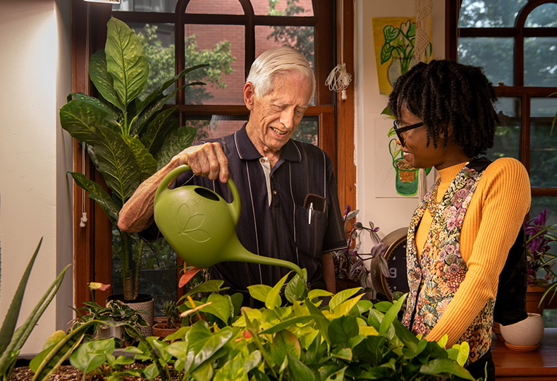 A resident is watering plants indoor with a water pot. A person is standing next to him. Both are smiling .
