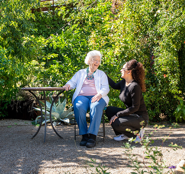 An associate is kneeling down laughing and smiling with a resident who is sitting down at a table outside.