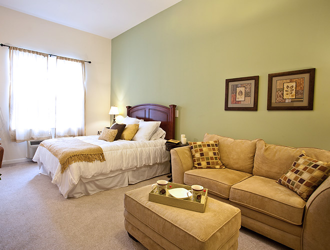 A bedroom with a light green wall, one queen size bed, tan sofa and tan ottoman.