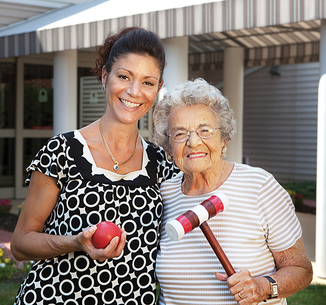 A person has arm around a resident. They are both smiling and holding a croquet set.