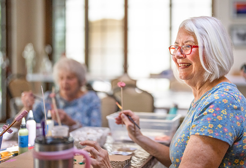 A resident is smiling and sitting at a table at an art class holding up a tool for painting.