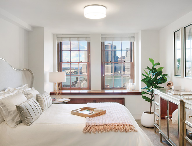 White bedroom with big bed, pillows, desk with chair by window with view of city.