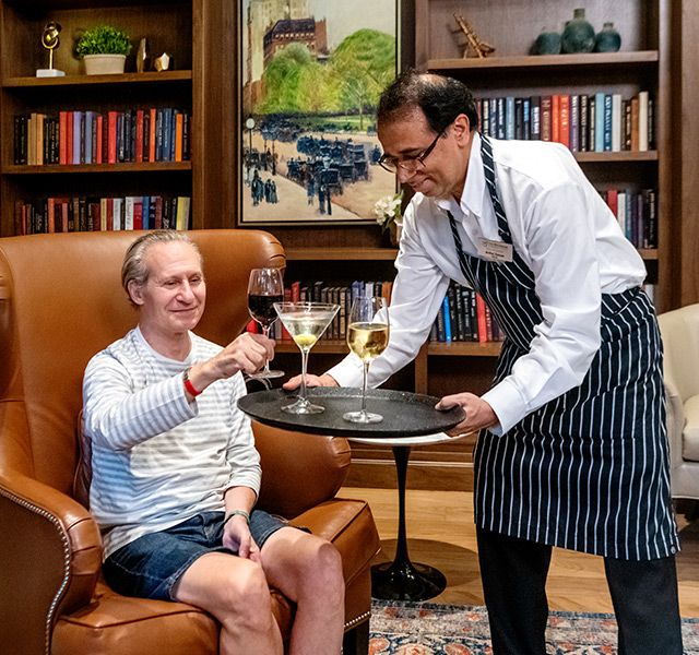 Waiter with tray serving glasses of wine with one resident sitting on a cozy leather arm chair reaching up for a glass.