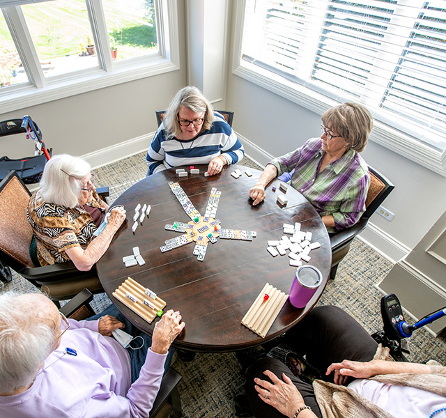 Multiple residents sitting around a round table playing a game of dominos.