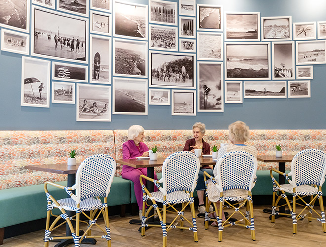 Three residents sitting at a table in front of a tall wall filled with framed black and white photos in a cafe.