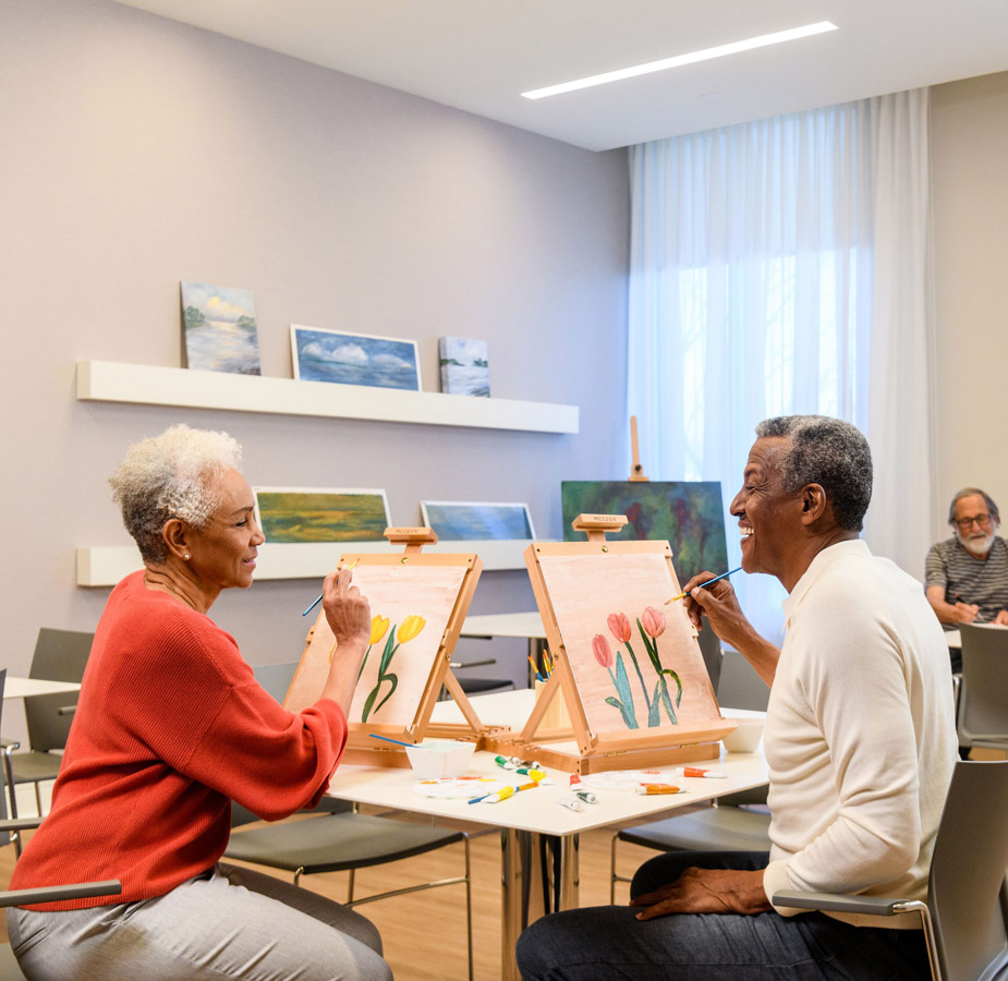 Two residents are painting on canvases in the art room.