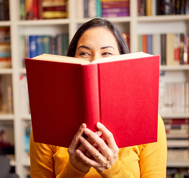 A person is holding a red covered book in front of their face.